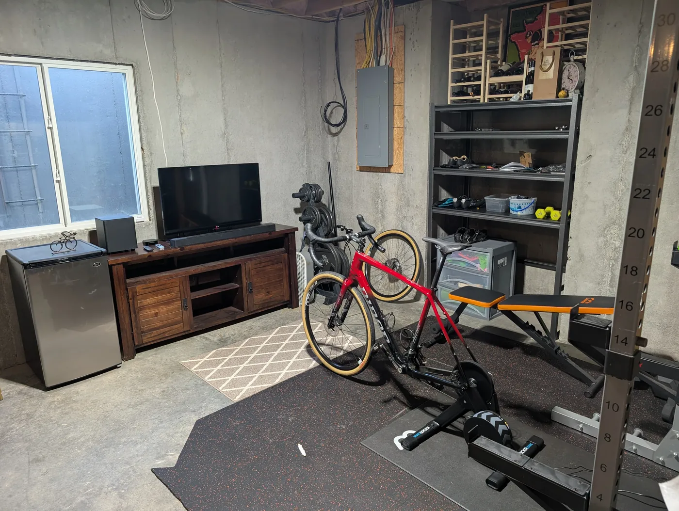 A finished basement training corner with a TV on a wood stand, a mini fridge, a weight rack and shelving, and a red gravel bike set up on a smart trainer