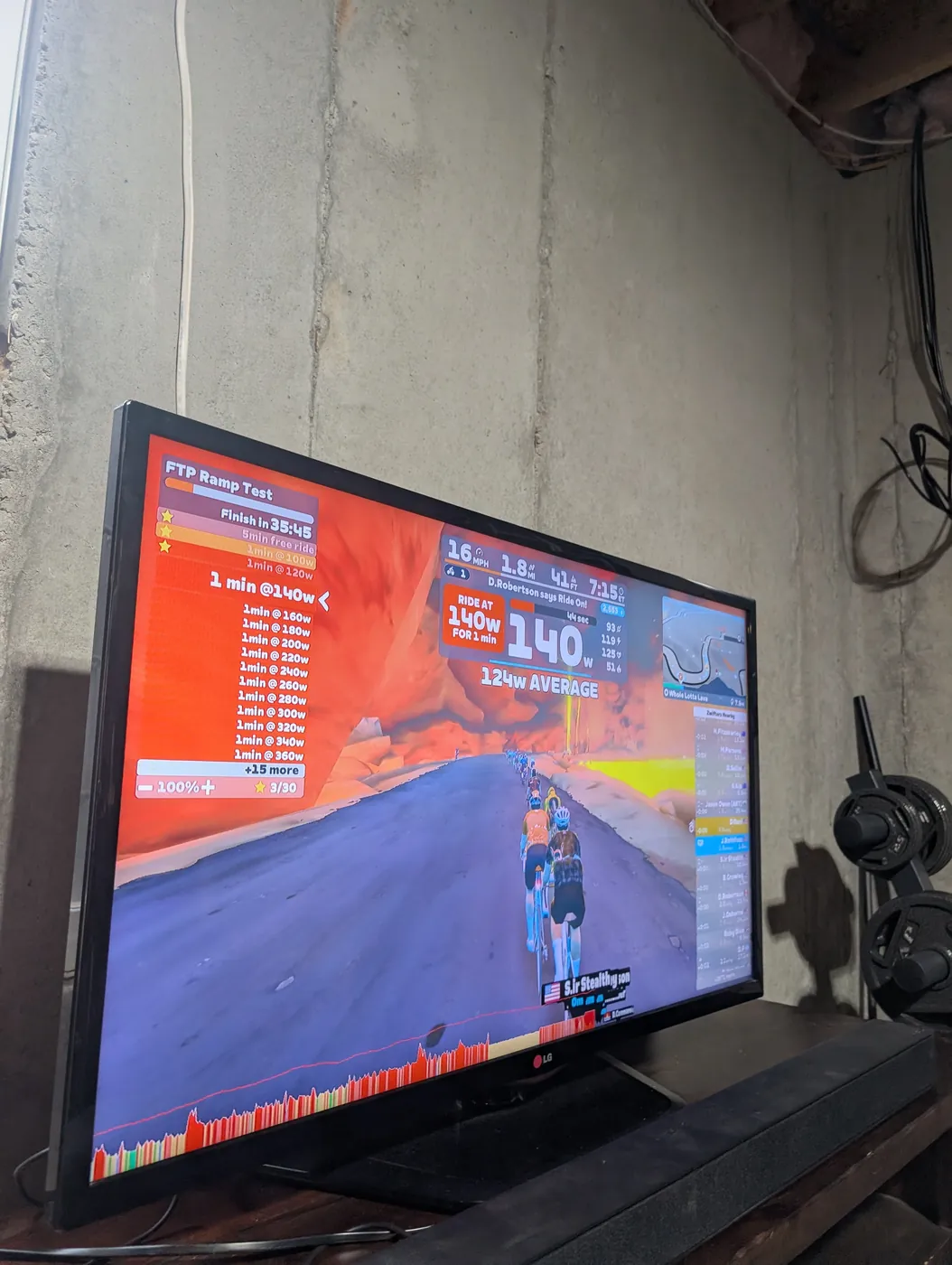 A TV screen showing a Zwift FTP ramp test, a line of riders climbing a road at 140 watts