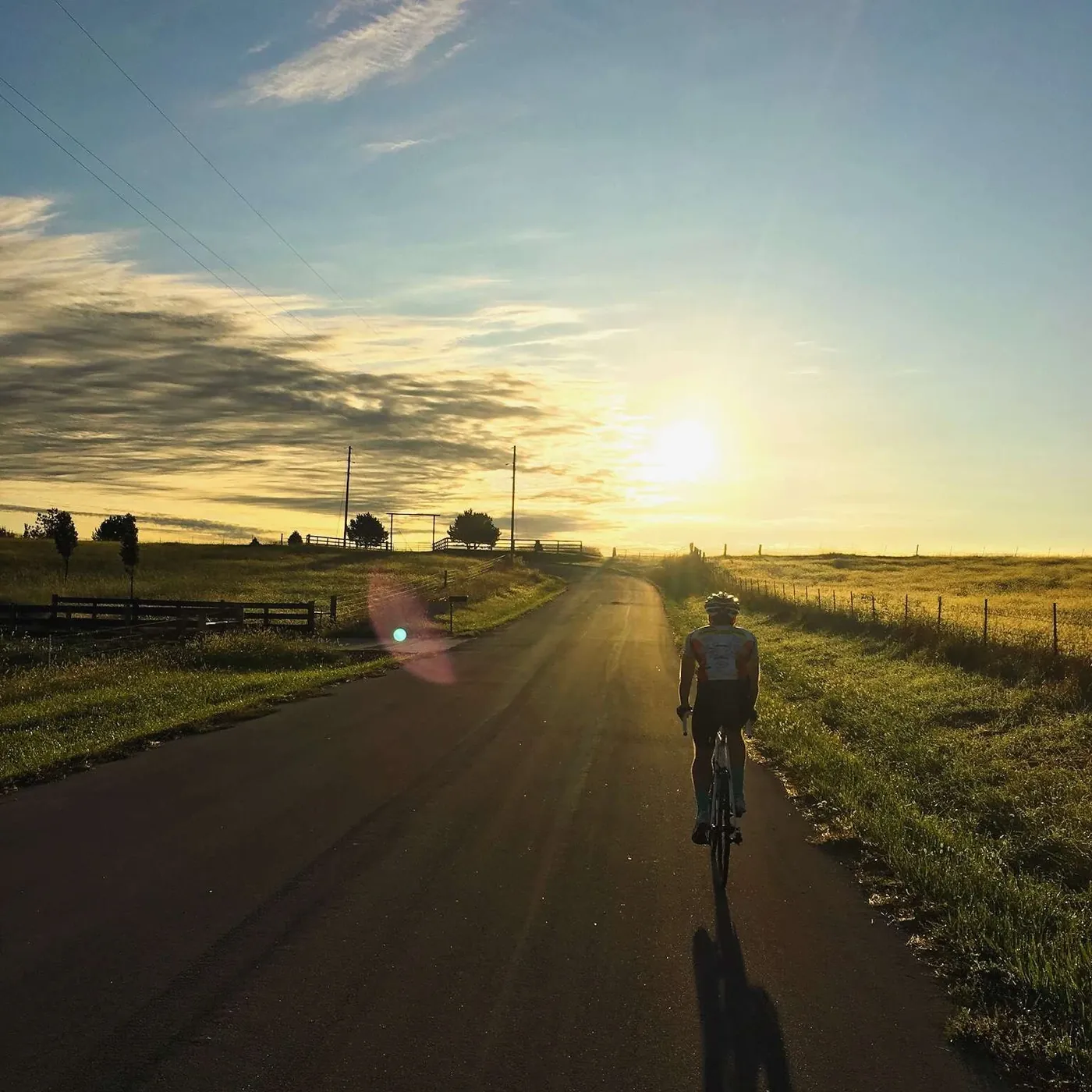 A cyclist riding away down an empty country road into a low sun, open fields and a fence line on both sides