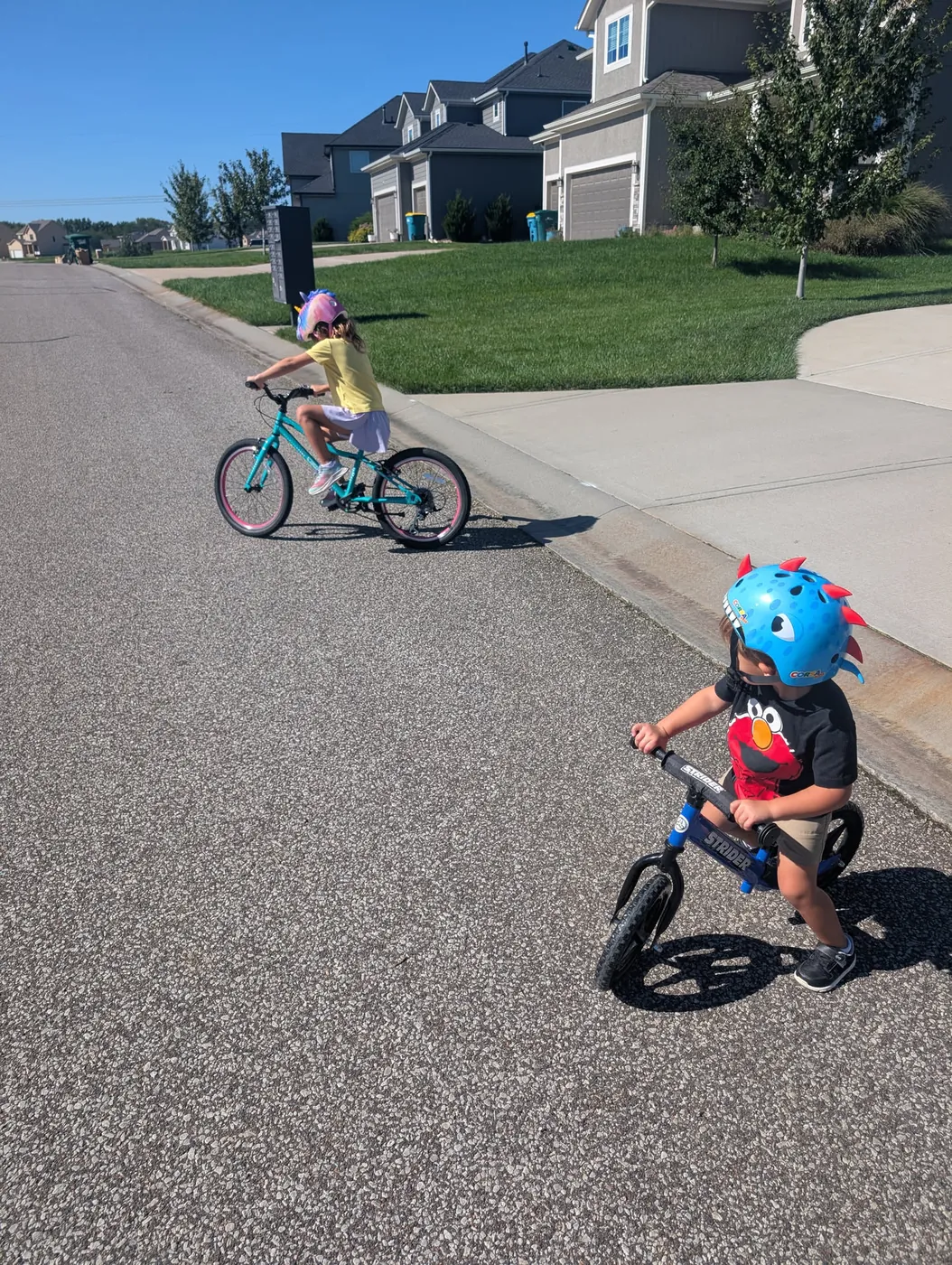 Two kids riding bikes down a suburban street, a girl on a teal bike ahead and a boy on a balance bike behind her