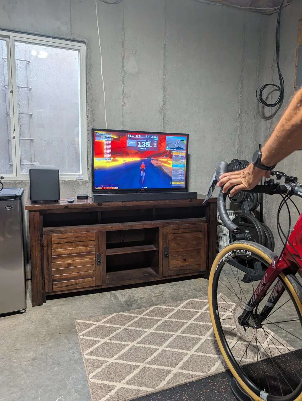 A man riding a bike set up on a smart trainer in a basement, hand on the shifter, a Zwift ride showing on the TV behind him
