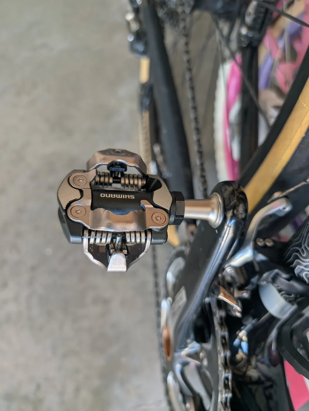 Close-up of a Shimano SPD clipless bike pedal mounted on a gravel bike crank, chain and rear wheel visible behind it