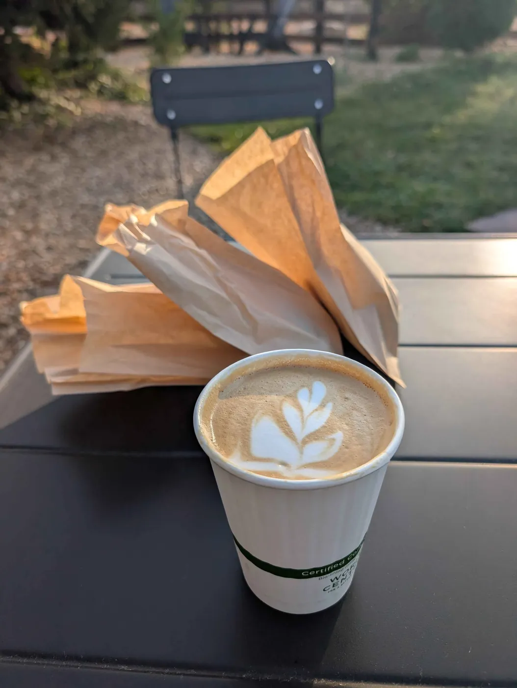 A latte with rosetta art in a paper cup on an outdoor table, empty pastry bags behind it