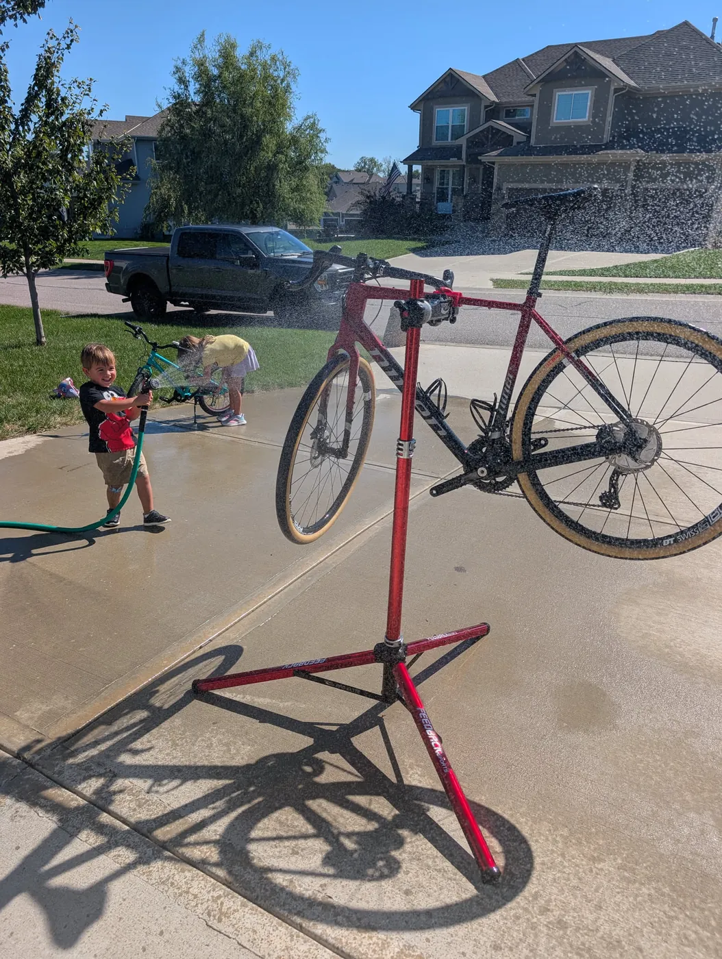 A closer view of the same gravel bike on its repair stand getting rinsed, the two kids still spraying their bikes behind it