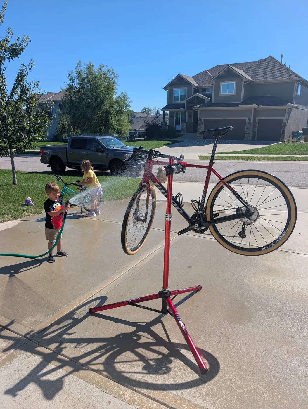 A red gravel bike on a repair stand being hosed down in a driveway, with two kids washing their own bikes with a hose in the background, a truck parked across the street
