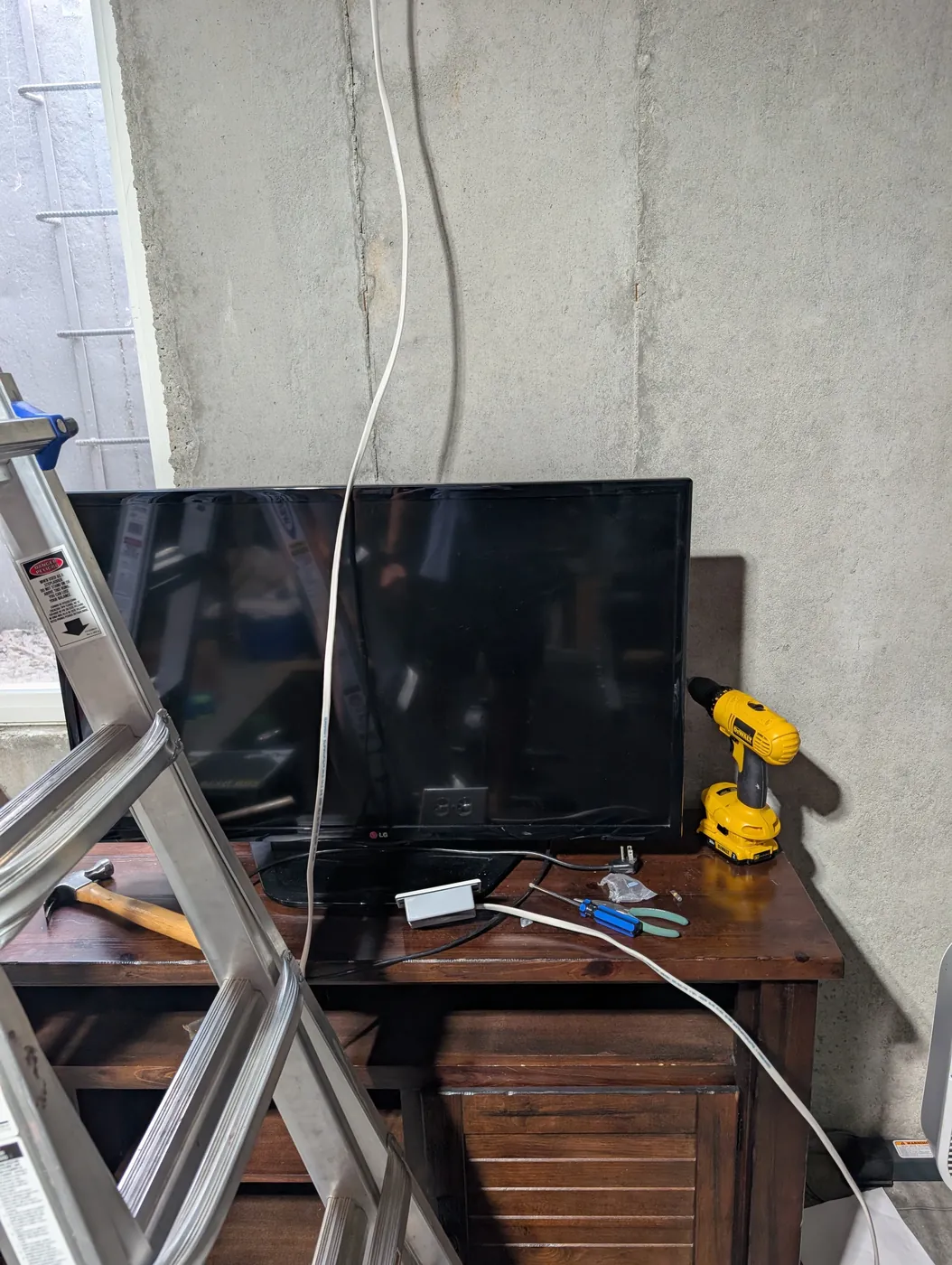 Two TVs sitting on a wood TV stand in an unfinished basement, cables running down the concrete wall, a drill and tools on top