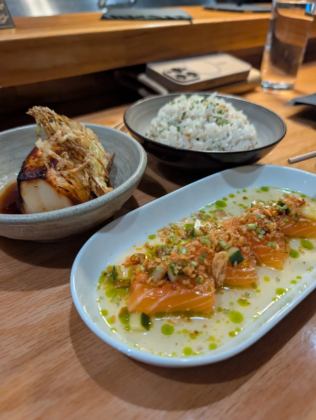 Three dishes on the bar: a piece of charred black cod under crispy fried onions, a bowl of rice, and salmon laid out in a pale sauce with green oil and chili crisp