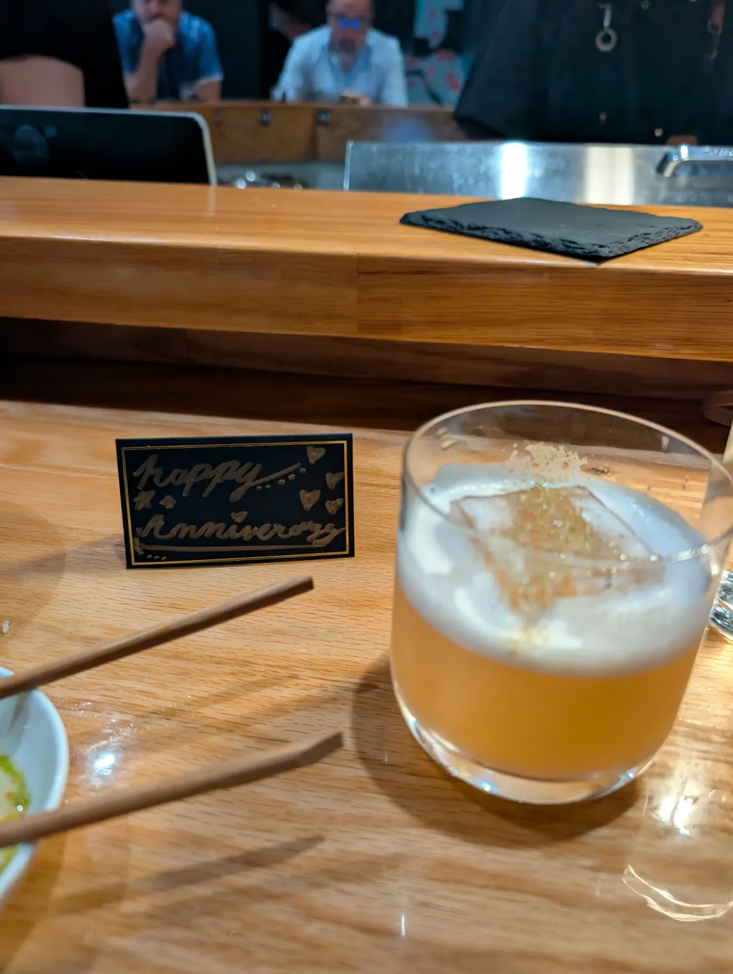 A foam topped cocktail on the bar next to a small black card reading happy anniversary in gold handwriting