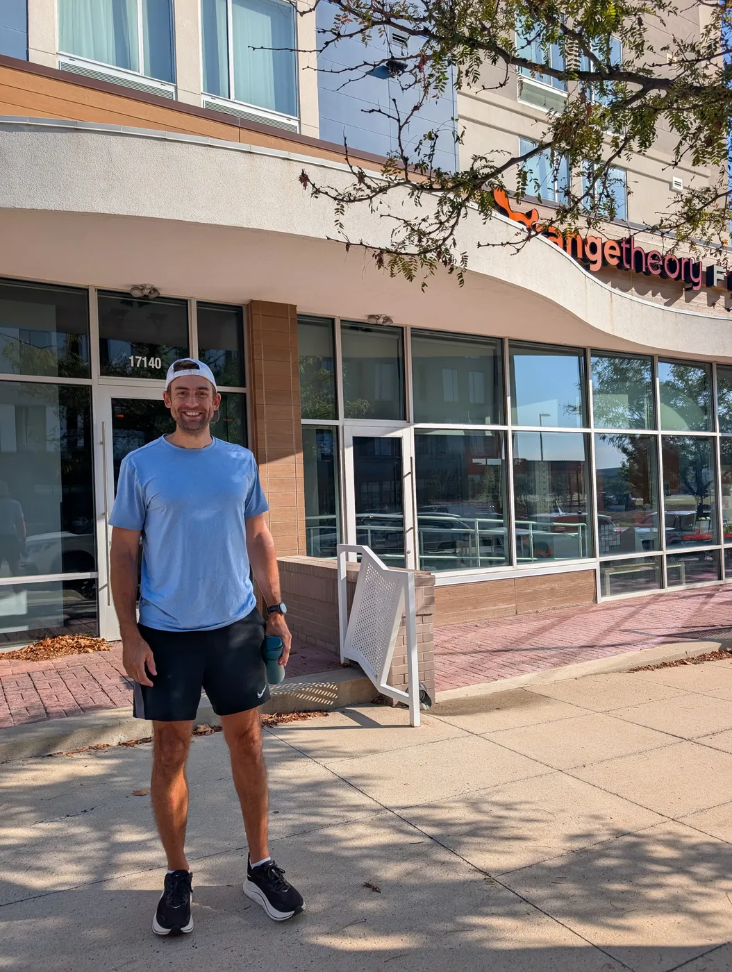 Me in a backwards cap, blue shirt and black shorts, standing on the sidewalk outside an Orangetheory Fitness with a water bottle