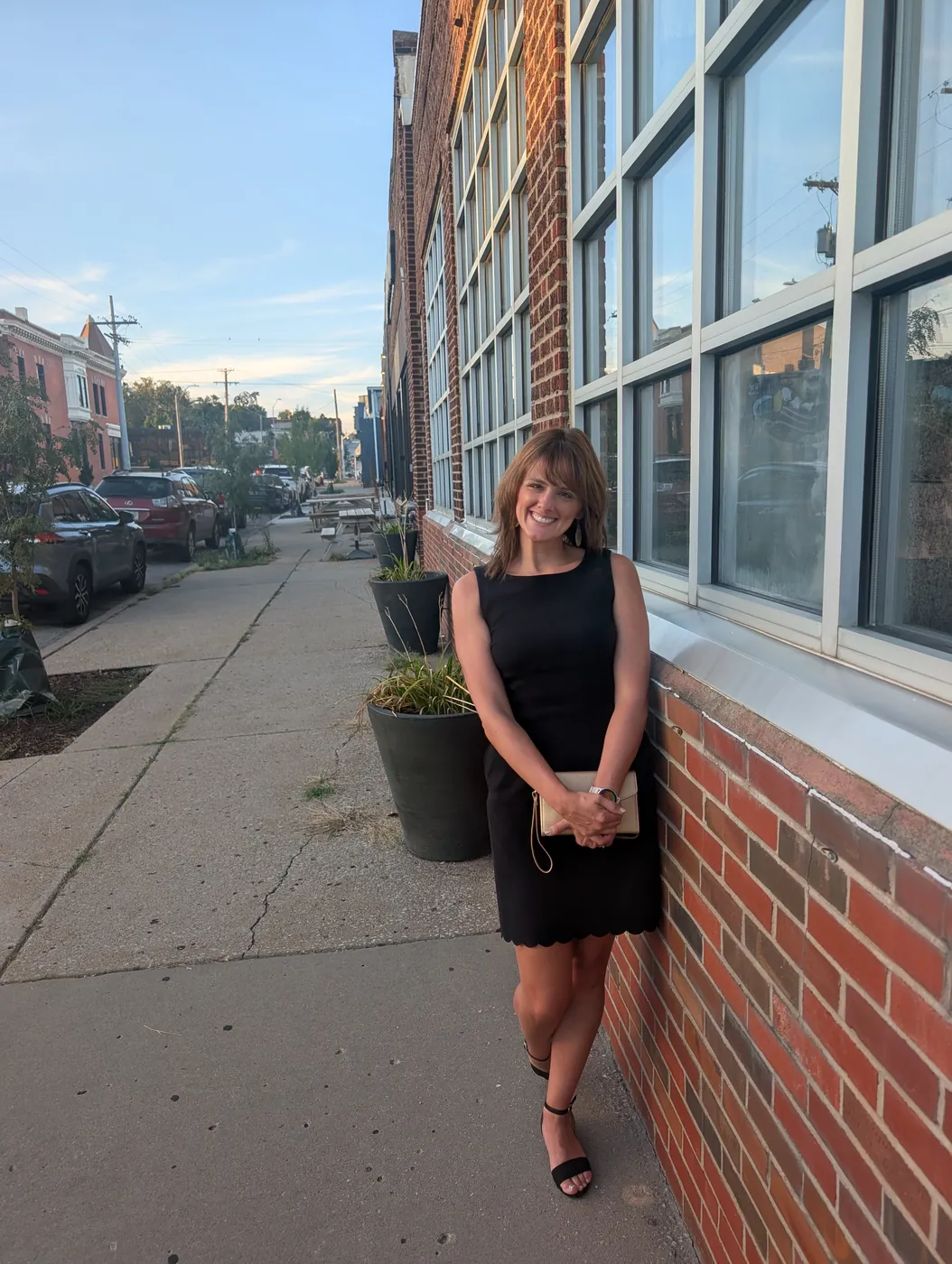 Natalie in a black dress leaning against the brick of an old building, sidewalk and parked cars behind her in the evening light