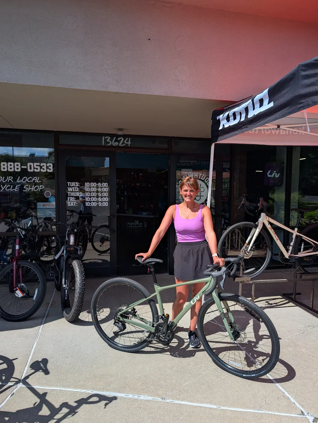 Natalie standing behind her new sage green Liv bike outside a bike shop, a black Kona tent set up by the door