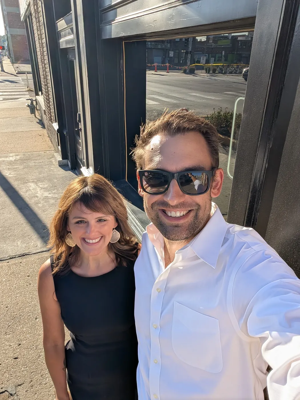 Natalie in a black dress and me in a white shirt and sunglasses, on the sidewalk outside a black storefront in the late sun