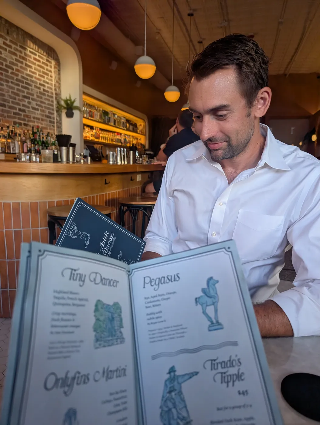 Me reading a tall cocktail menu at a bar, the back bar and warm globe lights behind me