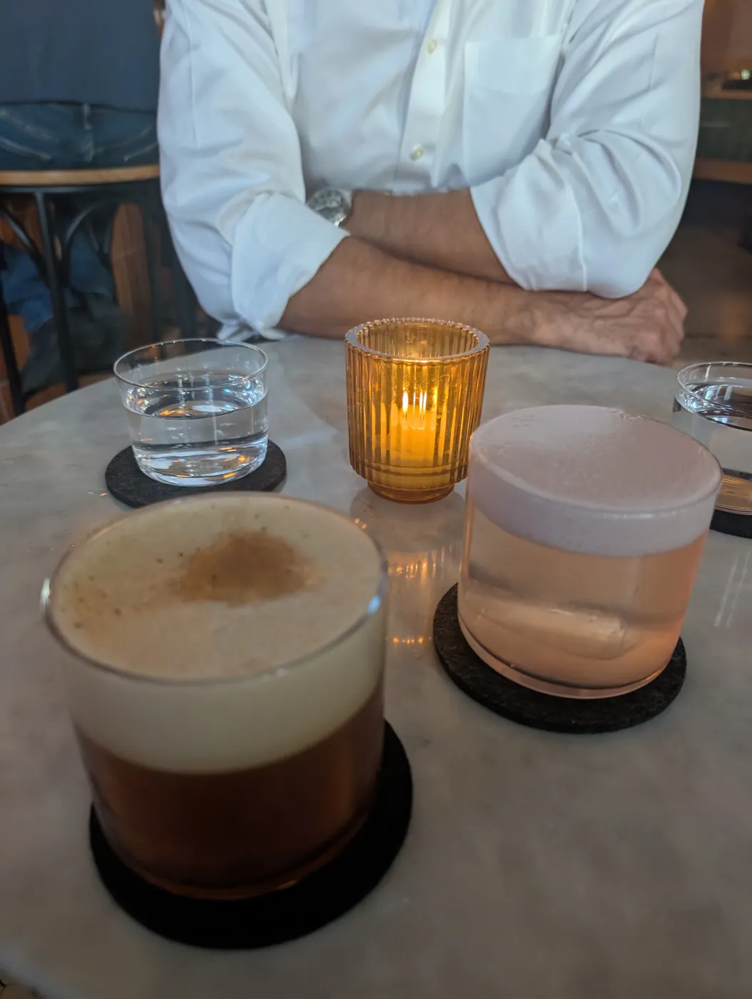 Two cocktails with thick foam tops on a marble table, an amber candle burning between them