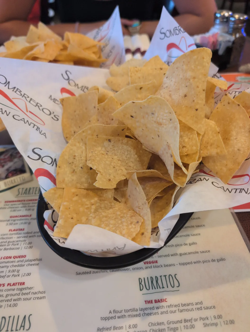 A black basket lined with printed paper and piled with tortilla chips, a menu on the table underneath