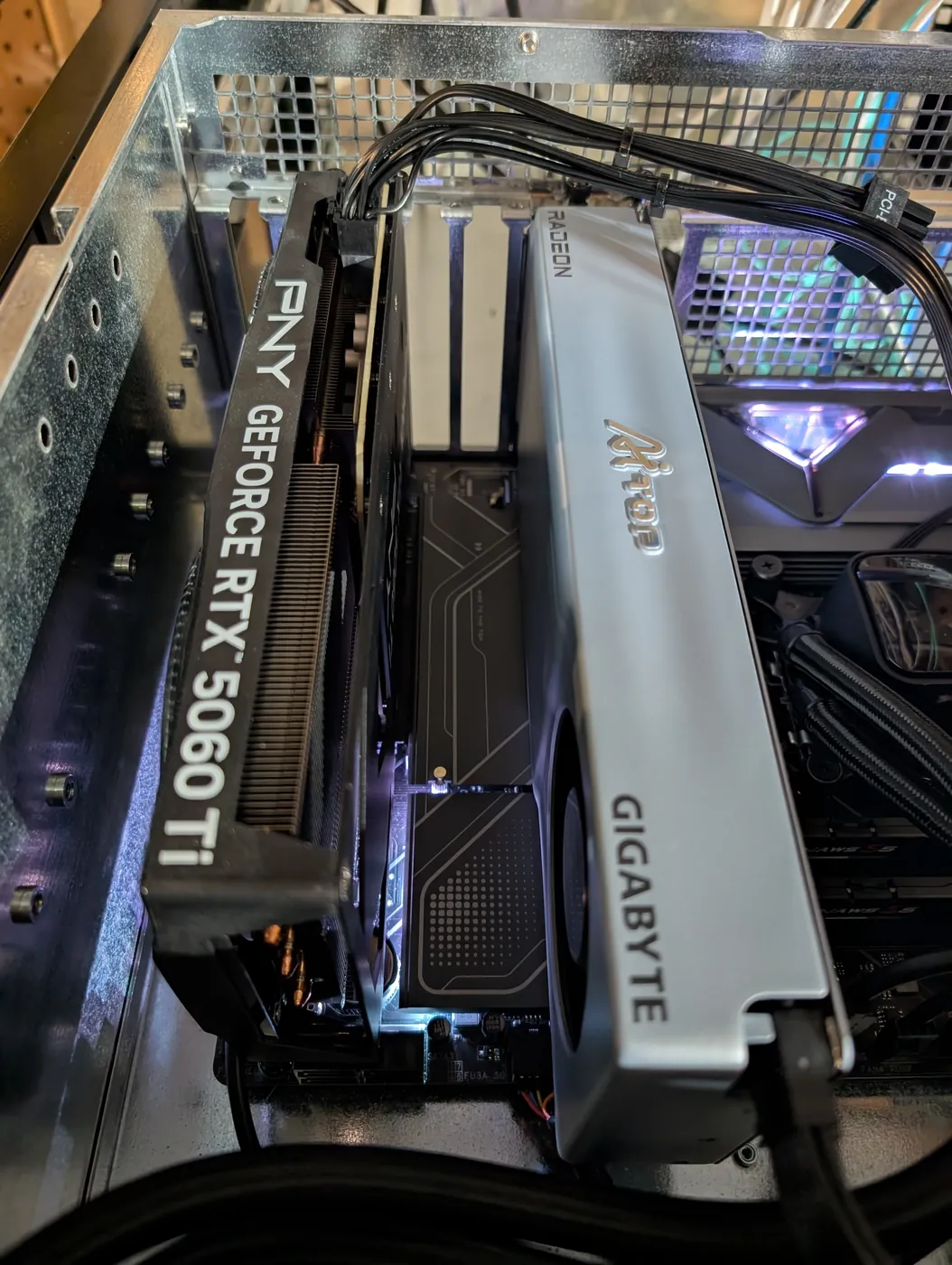 Looking down into an open computer case at two graphics cards side by side, a black PNY GeForce RTX 5060 Ti on the left and a silver Gigabyte Radeon card on the right