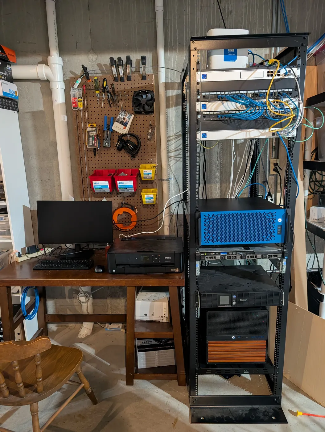 A black open-frame server rack in a basement next to a pegboard of tools and a wooden desk with a monitor and printer. The rack holds network switches with blue and yellow cables, a blue rackmount case, a thin one-unit server, a CyberPower battery backup and a black box with a wood-slat front