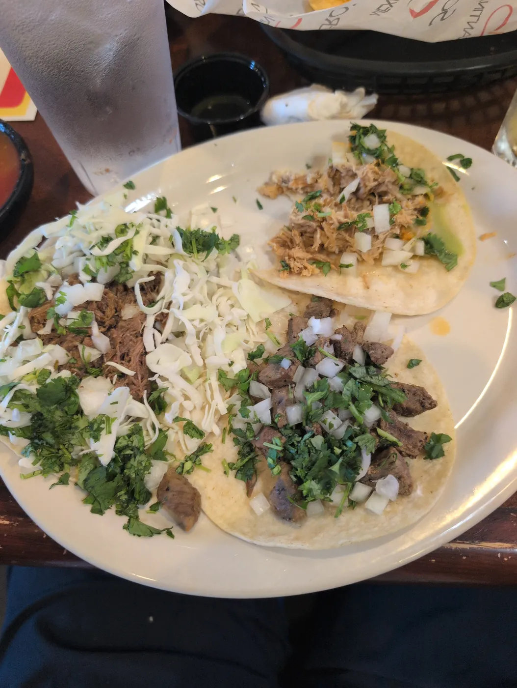 A white plate with three small tacos on corn tortillas, topped with shredded meat, cabbage, chopped onion and cilantro