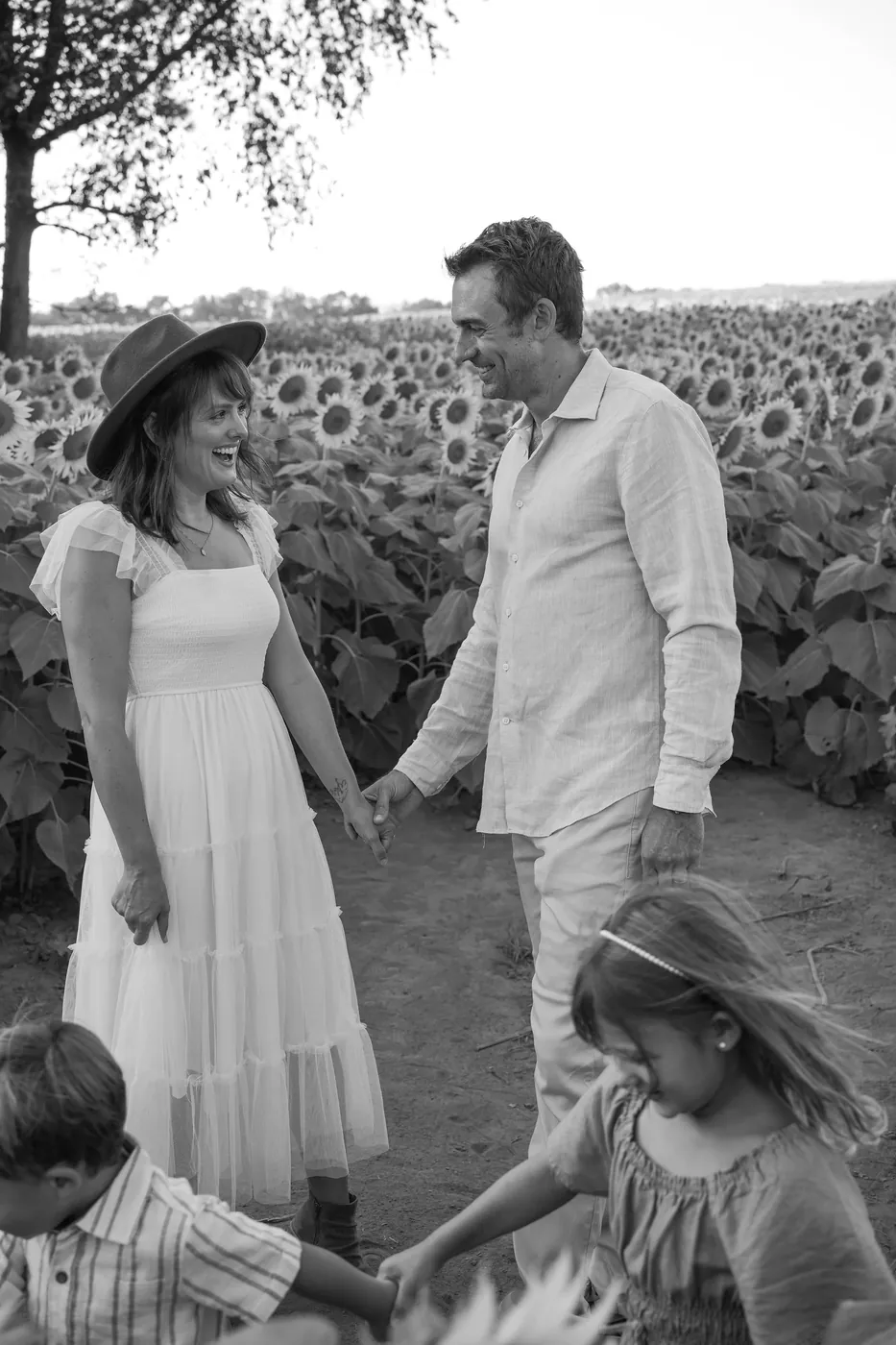 Black and white photo of John and Natalie holding hands at the edge of the sunflower field, laughing at each other, with their two kids blurred in the foreground