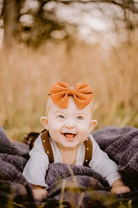A grinning baby propped on a blanket in a field, wearing an orange bow and brown suspenders