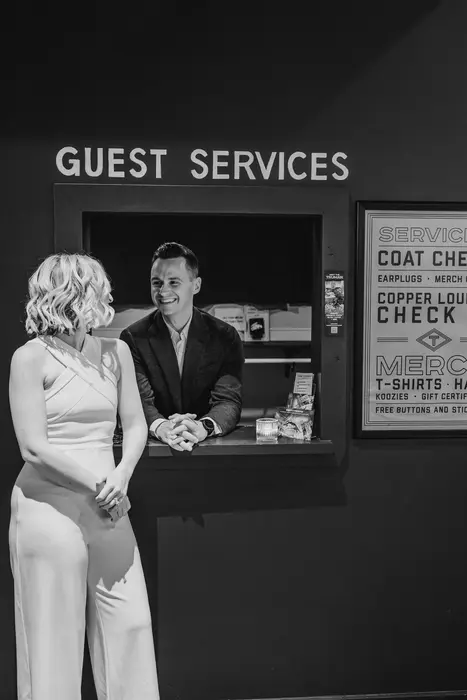 Black and white photograph of a bride in a white jumpsuit laughing with the groom, who leans through a venue guest services window