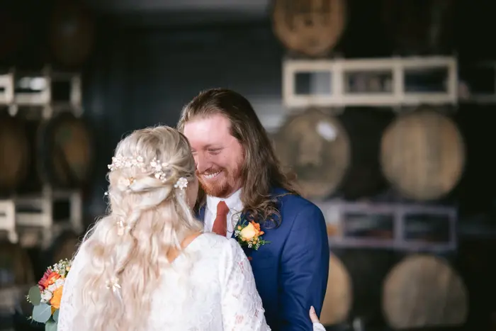 A bride and groom laughing together in front of stacked wine barrels, the groom in a blue suit with an orange boutonniere