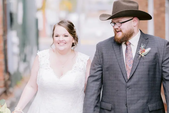 A bride and groom laughing as they walk hand in hand down a brick alley, the groom in a cowboy hat