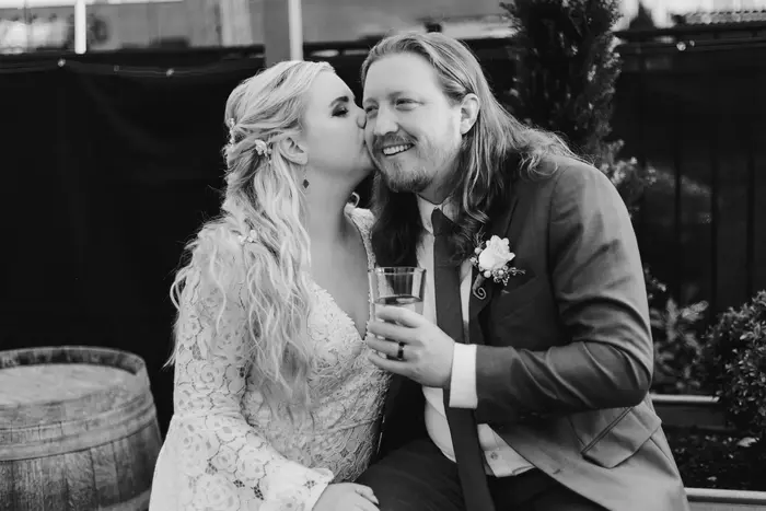 Black and white photograph of a bride kissing the groom on the cheek beside a wine barrel, a drink in his hand