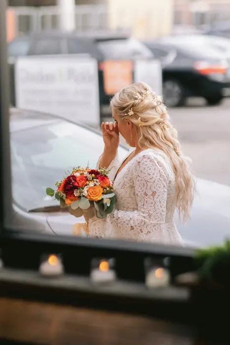 A bride seen from behind on a city street, holding an orange and red autumn bouquet, flowers braided into her hair