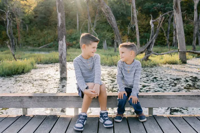 Two young brothers sitting on a boardwalk railing above a lily pond, laughing at each other in late afternoon light