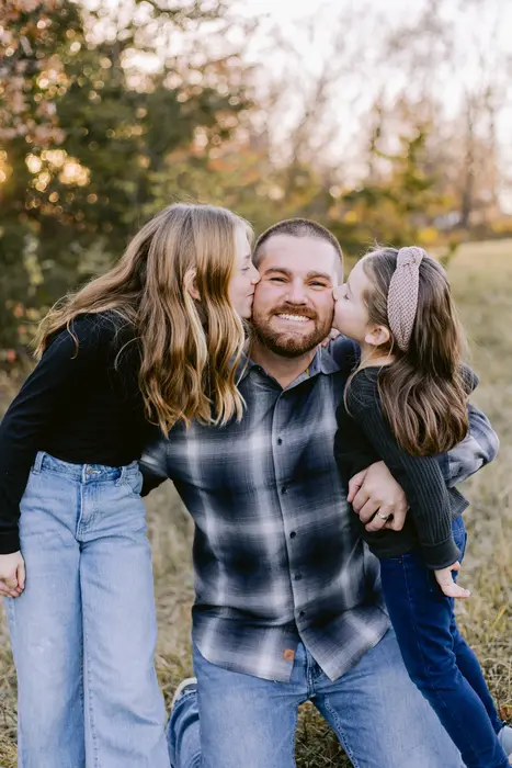 Two daughters kissing their laughing father on both cheeks in an autumn field