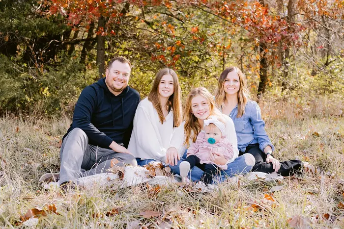 A family of five sitting together in fallen leaves beneath autumn trees