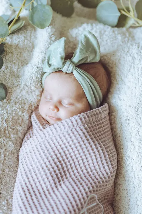 A sleeping newborn wrapped in a pink waffle swaddle with a green bow headband, eucalyptus sprigs alongside