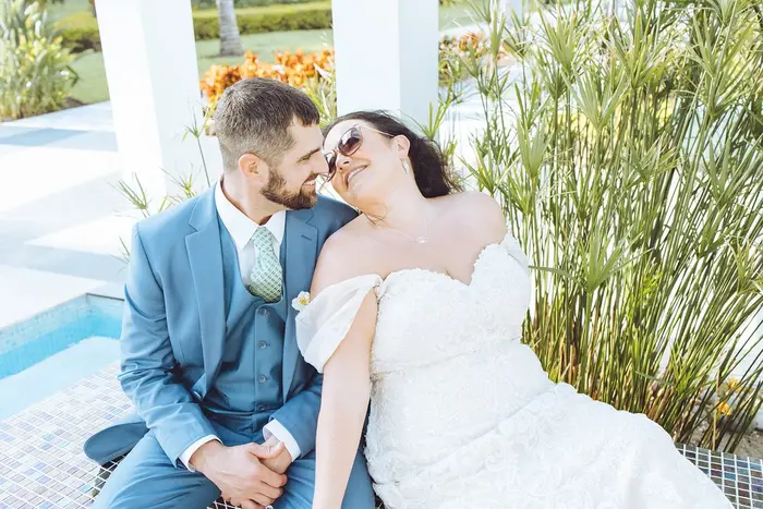 Newlyweds laughing together on a poolside bench at a tropical resort, the groom in a light blue suit