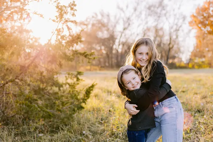 Two sisters hugging and laughing in a backlit autumn field at golden hour