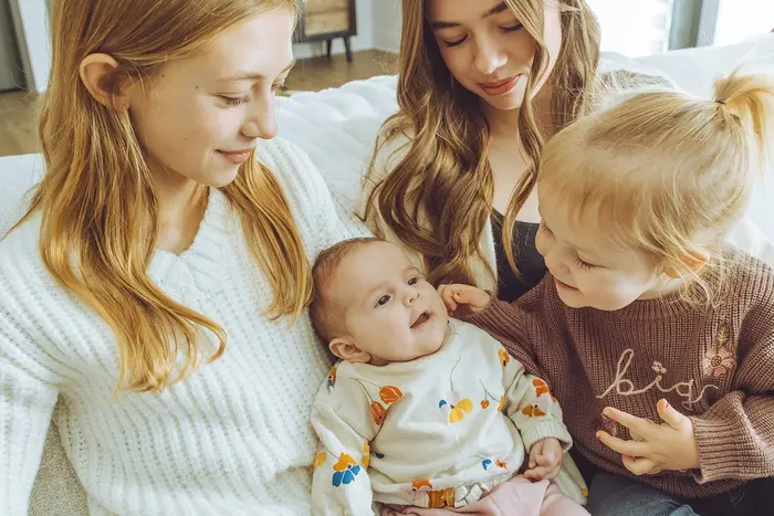 Three sisters gathered around a smiling baby on a couch in soft window light