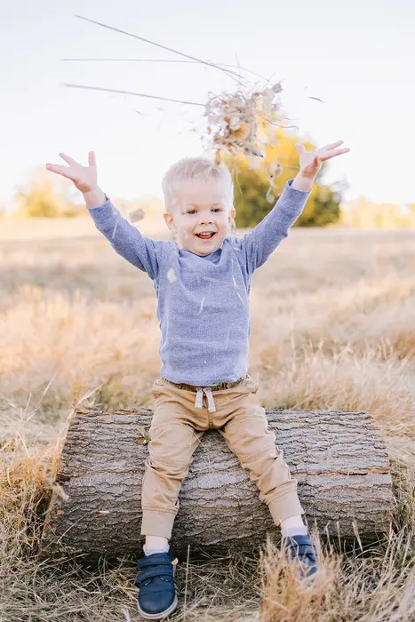 A laughing toddler sitting on a fallen log, throwing a handful of dried grass over his head