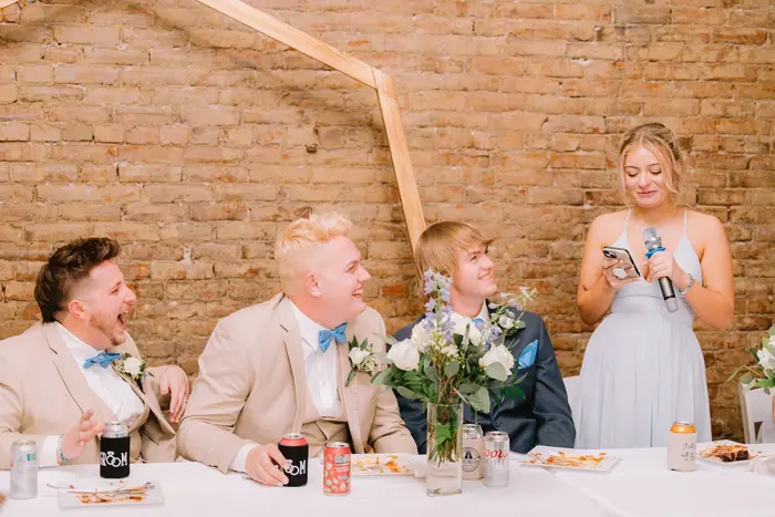 A bridesmaid reading a toast from her phone while the groom and groomsmen laugh at the head table, exposed brick behind them